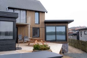 Casa moderna sul lungolago rivestita con pannelli WoodUpp: doghe verticali in legno chiaro per la facciata e decking WoodUpp sul terrazzo panoramico. Si vedono ampie finestre scorrevoli nere, tavolo picnic in legno, sedie pieghevoli e una fioriera vicino alla balaustra.