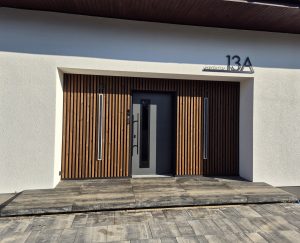 Ingresso moderno con rivestimento WoodUpp: pannelli in legno a listelli verticali scuro ai lati della porta grigia, due profili luminosi verticali integrati e maniglia nera. Il nome WoodUpp evidenzia il design a lamelle, creando contrasto con intonaco bianco e pavimento in pietra.