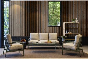 Interno soggiorno con parete rivestita dai pannelli a listelli in legno WoodUpp, finitura naturale con distanziatori scuri, e una scaffalatura aperta in legno WoodUpp a più ripiani; davanti divano e poltrone imbottite, tavolino basso con bicchieri e finestra che incornicia il verde esterno.