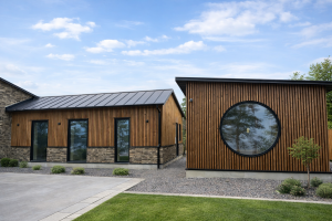 Esterno di edifici moderni rivestiti con eleganti pannelli a doghe di legno WoodUpp. La finitura in legno caldo crea un contrasto sorprendente con le cornici nere delle finestre rettangolari e della grande finestra circolare, oltre che con la base in pietra testurizzata.