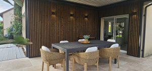 Veranda arredata con parete rivestita WoodUpp a doghe verticali in legno scuro con cornice nera, illuminazione da parete, e pavimento esterno in doghe grigie WoodUpp; tavolo da pranzo scuro e sei sedie in fibra naturale, vaso decorativo e piante a sinistra.