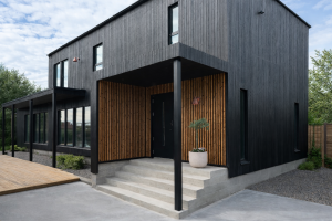 Esterno di una casa moderna con rivestimento scuro. L'ingresso rientrante è evidenziato dai pannelli a doghe verticali di WoodUpp in tonalità legno naturale caldo. Questo rivestimento WoodUpp crea un contrasto accogliente con la facciata nera circostante, sopra i gradini di cemento e la pianta in vaso.