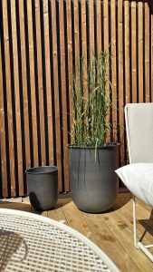 Schermatura verticale a listelli in legno WoodUpp dietro due vasi grigio scuro (uno grande con erba ornamentale, uno più piccolo vuoto), pavimento a doghe in legno WoodUpp, tavolo metallico bianco e sedia da esterno chiara con cuscino, illuminati da luce solare.