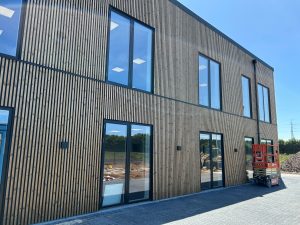 Facciata moderna rivestita da WoodUpp con doghe verticali in legno naturale, listelli sottili separati da giunti neri, grandi vetrate a doppio piano e porte a vetro. Sul piazzale si vede una piattaforma elevatrice rossa usata in cantiere e pavimentazione in autobloccanti.