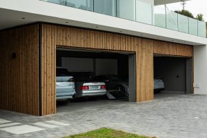 Facciata garage moderna con rivestimento WoodUpp: listelli di legno verticali naturali e regolari che coprono due grandi portoni del box auto. Si vedono tre aperture parzialmente aperte, pavimentazione in pietra, automobili parcheggiate e balcone superiore con parapetto in vetro, con finiture metalliche scure.