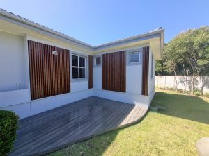 Vista di una casa bianca con rivestimento esterno WoodUpp: pannelli verticali di listelli in legno scuro installati ai lati delle finestre e un decking WoodUpp grigio a profilo curvo. Giardino erboso e recinzione chiara sullo sfondo, ben rifinito sotto cielo azzurro.