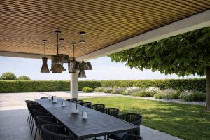 Un elegante patio esterno presenta un soffitto rivestito con i pannelli acustici a listelli di legno chiaro di WoodUpp, che donano calore e stile moderno. L'area pranzo sottostante include un lungo tavolo scuro, sedie intrecciate nere e lampade a sospensione, con vista su un giardino curato.