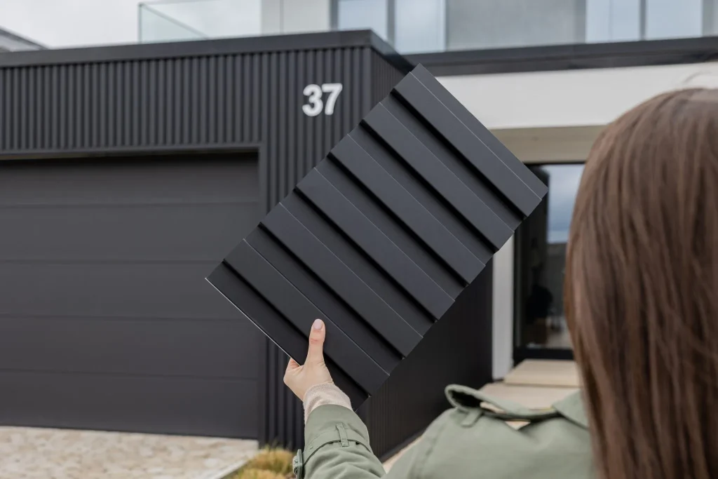 Una persona impugna un campione di pannello WoodUpp nero rigato con listelli rettangolari, fronte a una casa moderna rivestita con pannelli WoodUpp neri a stecche verticali e portone garage scuro. Il campione evidenzia profili modulari, profondità delle incisioni e finitura opaca.