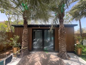 Edificio da giardino rivestito con pannelli verticali in legno WoodUpp, portafinestra scorrevole nera a vetro centrale, due palme davanti, luci a sospensione lungo il tetto e vasi dorati ai lati. Si vedono listelli in legno WoodUpp e finitura nera del telaio.