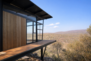 Vista esterna di una cabina moderna con rivestimento verticale in legno di WoodUpp sulla parete. I listelli di legno caldo contrastano con la struttura in acciaio nero e le grandi vetrate. Una terrazza in legno si affaccia su un vasto paesaggio collinare e secco sotto un cielo blu.