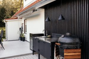 Area cucina esterna WoodUpp con mobile contenitore nero su gambe e piano inox con lavello, frigorifero compatto, lampade sospese nere e barbecue kamado WoodUpp nero con ripiani in legno; parete rivestita in doghe di legno scuro, terrazza in gres e dettagli minimal.