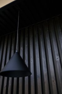 Interno con rivestimento WoodUpp a listelli di legno verniciato nero disposti verticalmente, accento di venature e viti a vista; lampada a sospensione WoodUpp dal paralume conico nero appesa in primo piano, filo sottile e finitura opaca, atmosfera moderna e minimale.