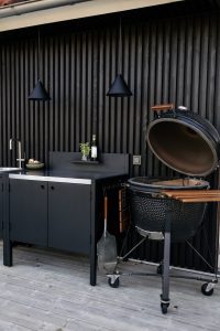 Area barbecue WoodUpp: mobile cucina esterno nero con lavabo, piano in acciaio, ante contenitive e sottile mensola con bottiglia ed erba aromatica; due lampade a sospensione coniche nere WoodUpp; barbecue kamado nero su carrello con maniglie e ripiano laterale in legno.