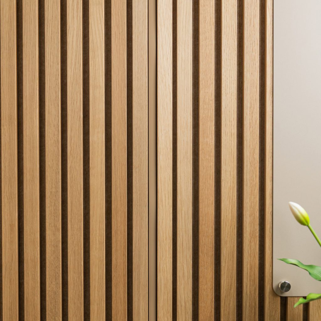 Superficie di pannelli dogati WoodUpp in legno chiaro con listelli verticali regolari e scanalature scure tra le doghe; a destra si vede una piastra metallica fissata con una vite e, in basso a destra, una gemma di fiore bianco con foglia verde.