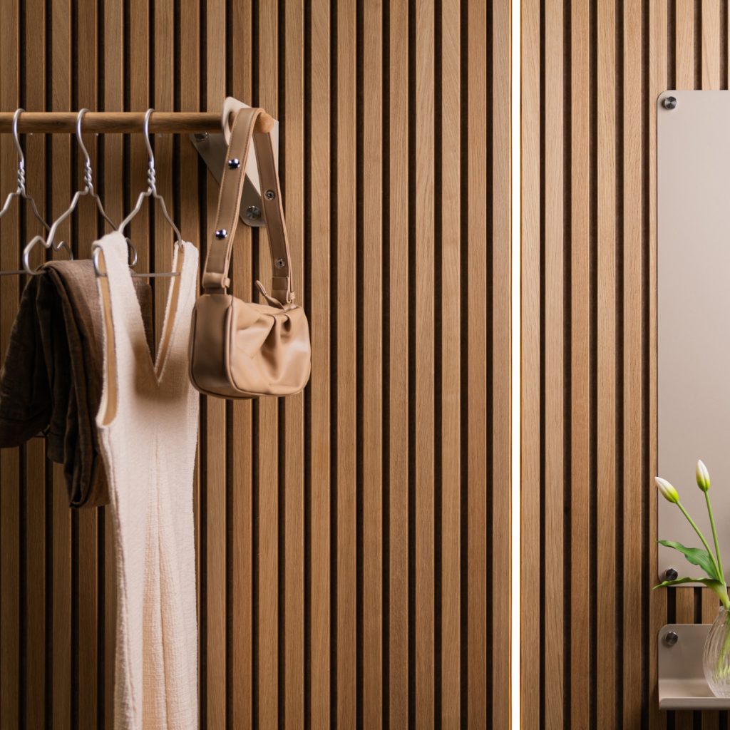 Pannello dogato in legno WoodUpp a listelli verticali chiari; prodotti WoodUpp visibili: barra appendiabiti in legno con staffe metalliche che sostiene grucce, un abito e una borsa beige. A destra mensola con specchio e vaso con fiori, illuminazione sottile tra i listelli.