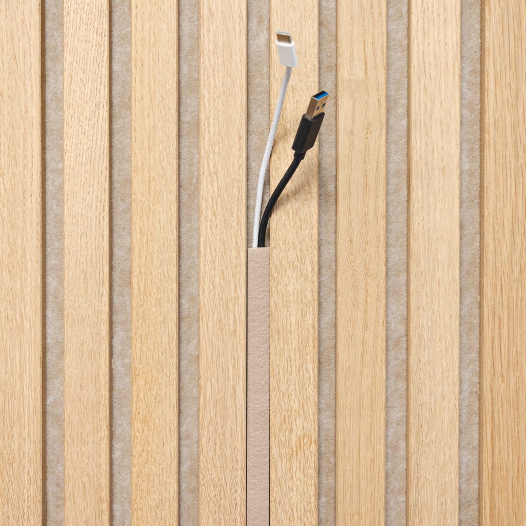 Primo piano di un pannello acustico WoodUpp a doghe in legno chiaro con intercapedine in feltro beige; al centro si nota una canalina passacavi WoodUpp rettangolare che lascia uscire due cavi (bianco e nero). Texture del legno e disposizione regolare delle doghe sono evidenti.