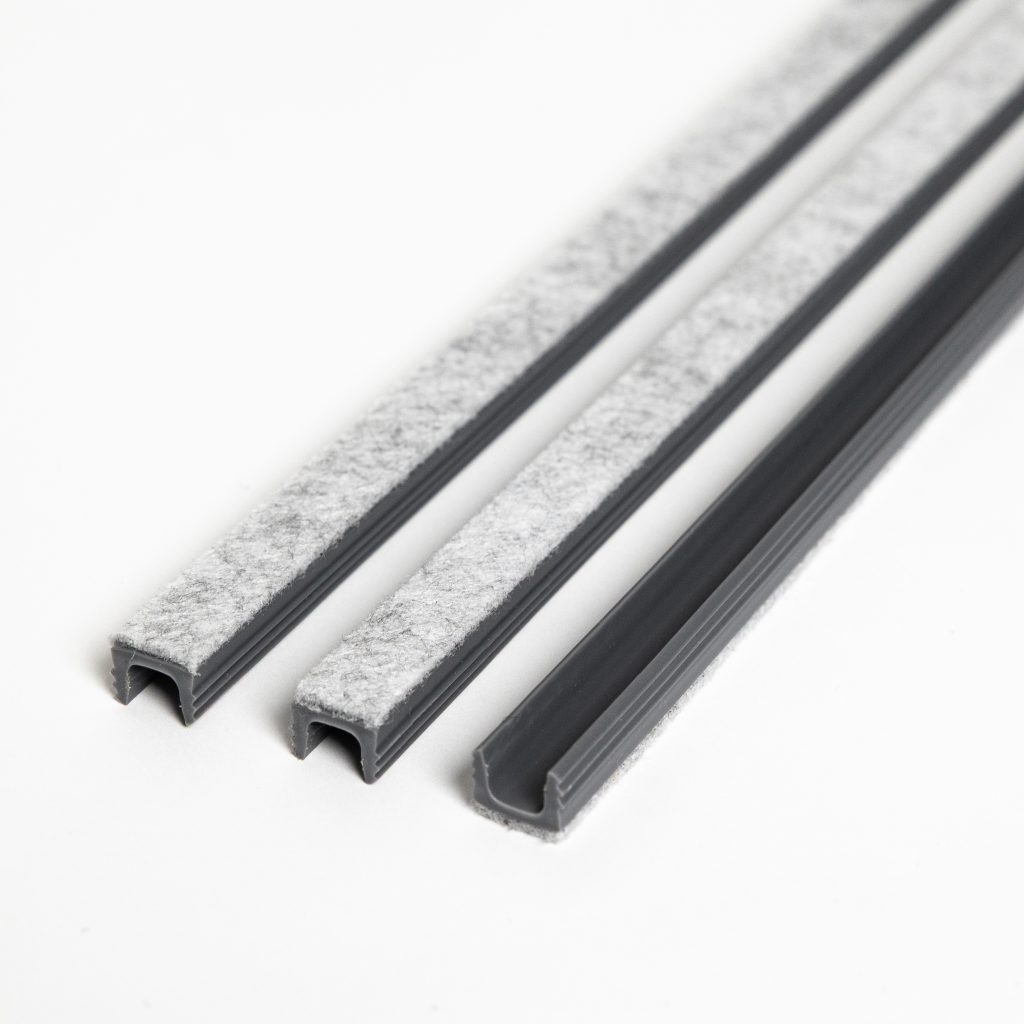 Immagine WoodUpp: primo piano di tre profili in plastica grigia a U con striscia di feltro grigio chiaro applicata sulla superficie, utilizzati come protezioni antiscivolo e anti-graffio per mobili e ante. Eccoli disposti su sfondo bianco, visualizzati in dettaglio, nitidi.