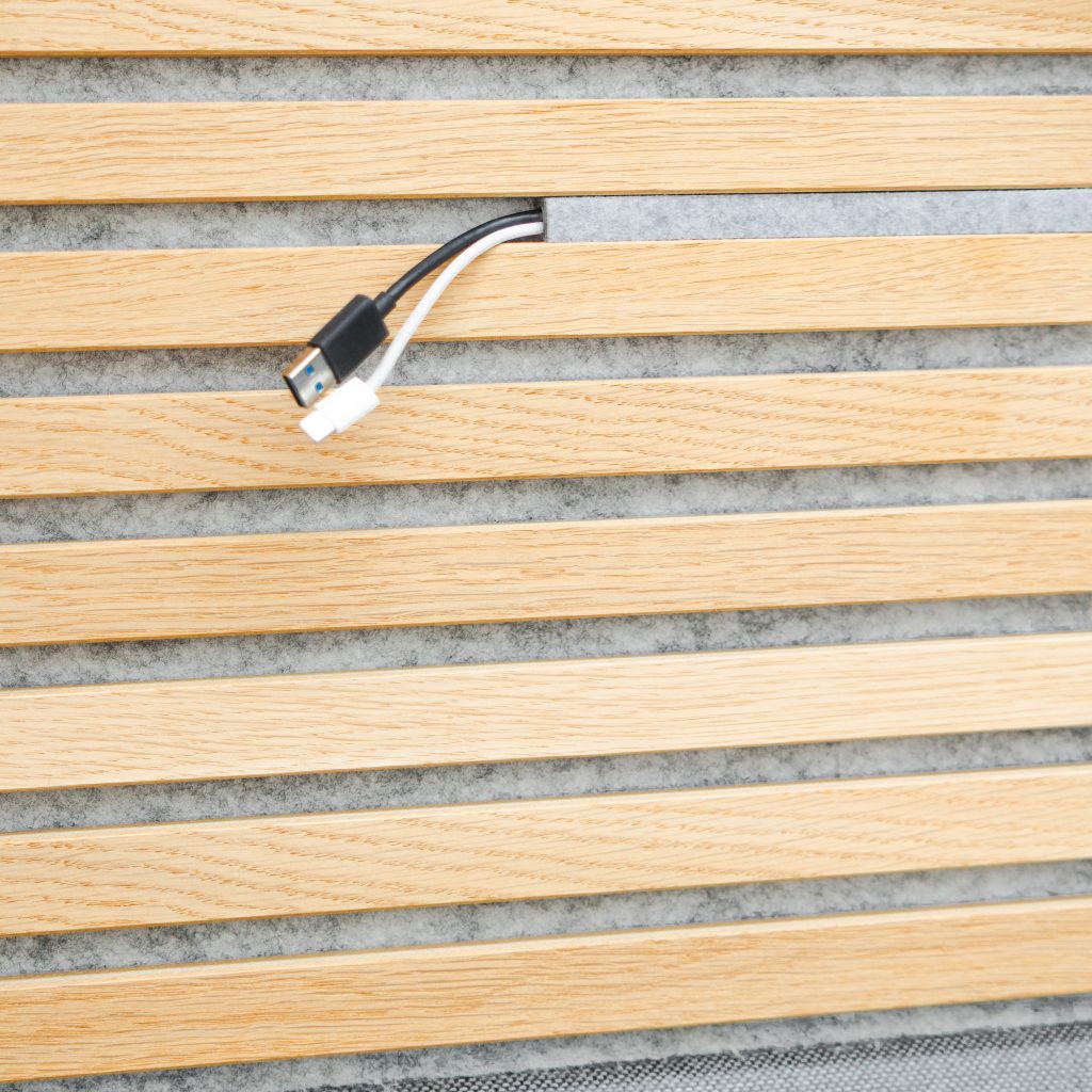Pannello WoodUpp a doghe orizzontali in rovere chiaro con venature naturali e inserto fonoassorbente in feltro grigio; apertura passacavi centrale rifinita da cui escono due cavi di ricarica (USB-A nero e cavo bianco). Design modulare WoodUpp, finitura liscia e interspazi regolari.