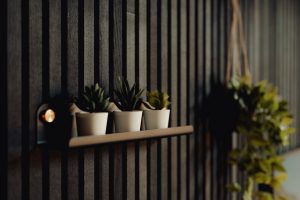 Parete a listelli verticali WoodUpp in legno scuro con mensola sottile WoodUpp montata, tre vasetti bianchi con succulente sulla mensola, una piccola luce calda laterale e una pianta sospesa sfocata sul lato destro, dal design moderno e minimal che valorizzano l'ambiente.