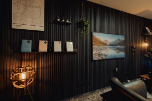 Interno moderno con parete WoodUpp a listelli di legno scuro che copre tutta la parete; mensole sospese WoodUpp nere tengono libri e piccoli vasi. TV con paesaggio montano, lampada da terra a gabbia, poltrona in pelle e tappeto shag completano l'ambiente caldo e accogliente.