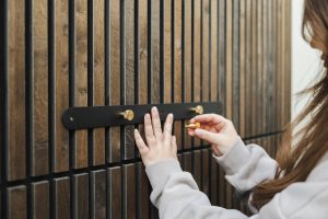 Primo piano su parete rivestita WoodUpp in listelli di legno scuro verticali; una barra di montaggio nera WoodUpp con pomelli/appendini dorati in ottone viene fissata da una persona, le sue mani si vedono mentre avvitano un pomello, indossando una felpa grigia.