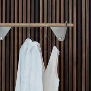 Dettaglio WoodUpp: parete a doghe in rovere scuro e chiaro, barra appendiabiti in legno massello con staffe metalliche triangolari color crema, due grucce metalliche con camicia bianca e top beige appesi, design minimal ed elegante, finiture naturali, perfetto per ingressi e cabine armadio.