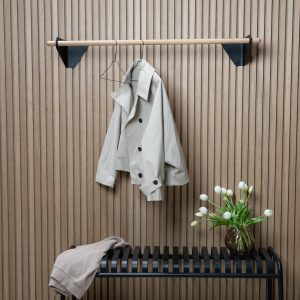 Parete rivestita in listelli di legno con attaccapanni WoodUpp: una barra cilindrica in legno sostenuta da staffe nere triangolari, e panca WoodUpp nera a listelli curvi. Sulla barra un soprabito beige appeso, sulla panca una felpa e un vaso di tulipani bianchi.
