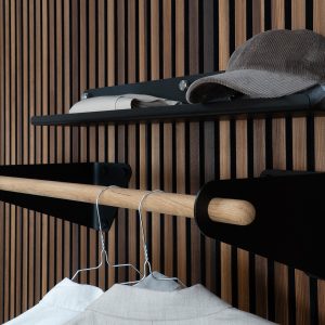 Pannello verticale a listelli in legno con sistema WoodUpp: mensola nera in metallo con viti a vista, asta appendiabiti in rovere massello sostenuta da staffe nere sagomate, due grucce metalliche con camicie appese e sopra ripiano con cappello e indumento piegato.
