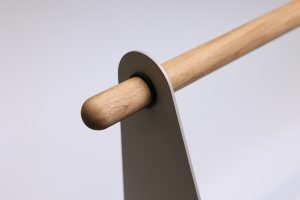 Primo piano di prodotti WoodUpp: un'asta in legno chiaro (rovere) cilindrica e levigata inserita in un supporto verticale minimalista in metallo grigio, con boccola nera in gomma tra legno e metallo; finitura naturale, linee moderne e assemblaggio curato e dal design elegante.