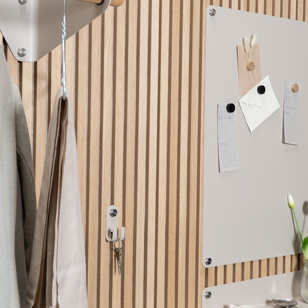 Parete WoodUpp in pannelli a doghe di legno chiaro con ganci e portachiavi da parete WoodUpp che reggono chiavi e capi appesi; pannelli organizer magnetici grigio chiaro fissati con distanziali metallici, note e magneti, e un piccolo vaso con tulipano bianco sul bordo.