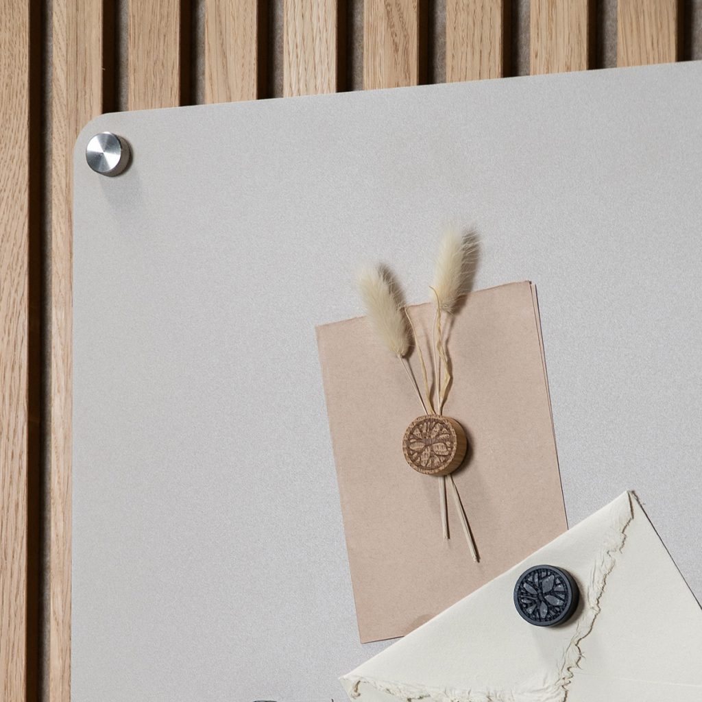 Immagine WoodUpp: pannello a lamelle in legno chiaro sullo sfondo, lavagna magnetica grigio chiaro fissata con distanziali metallici e due magneti rotondi WoodUpp — uno in legno inciso che trattiene biglietto con steli secchi, e uno scuro che fissa una busta.