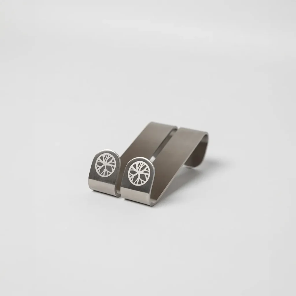 Immagine di WoodUpp che mostra due clip portafoglio in acciaio satinato, sagomate a molla e posizionate affiancate su fondo bianco. Le estremità riportano un logo circolare inciso a motivo radiale; design minimalista, finitura metallica e dettagli raffinati caratterizzano questi prodotti WoodUpp.