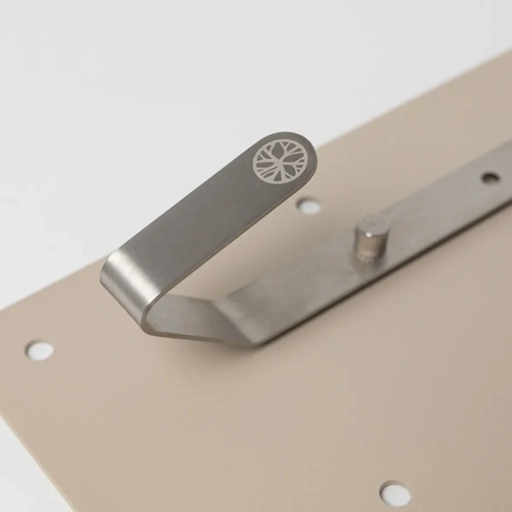 Primo piano di un prodotto WoodUpp: gancio appendiabiti in acciaio satinato con logo WoodUpp inciso a forma di sezione di tronco sul puntale, fissato a una piastra di montaggio color sabbia con fori per viti e perno di bloccaggio visibile.