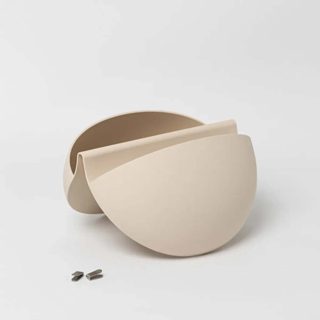 Immagine di WoodUpp che mostra un vaso/portapiante da parete color sabbia in due gusci curvi sovrapposti, forma semicircolare liscia con apertura frontale per piante; design modulare minimalista in materiale opaco. Accanto si vedono due piccole clip metalliche per il fissaggio a parete.