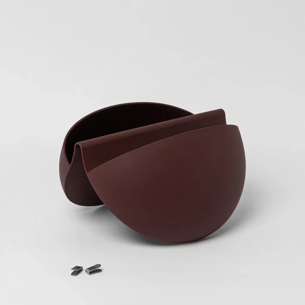 Immagine di un prodotto WoodUpp: due gusci curvi in metallo marrone scuro opaco sovrapposti a formare un portaoggetti o vaso basso dal design organico e minimalista dall'aspetto elegante, con una fessura centrale. Davanti tre piccole clip metalliche separate, su sfondo chiaro.