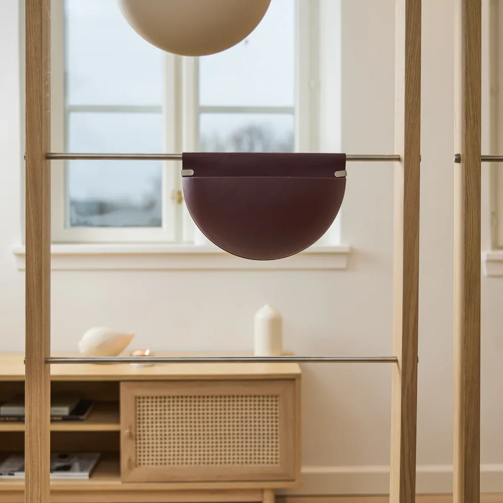 Immagine WoodUpp di una struttura divisoria in rovere chiaro con barre metalliche; appesa una tasca portaoggetti semicircolare in pelle bordeaux fissata con clip metalliche. Sullo sfondo mobile basso in legno con anta a intreccio, finestra, candela e oggetti decorativi, atmosfera minimal e naturale.