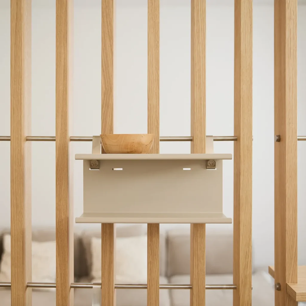 Immagine WoodUpp: parete divisoria modulare in legno rovere chiaro con doghe verticali e barre metalliche, con una mensola modulare beige WoodUpp fissata al pannello. Sulla mensola è appoggiata una ciotola in legno chiaro; stile minimal, finiture naturali e montaggio a clip.