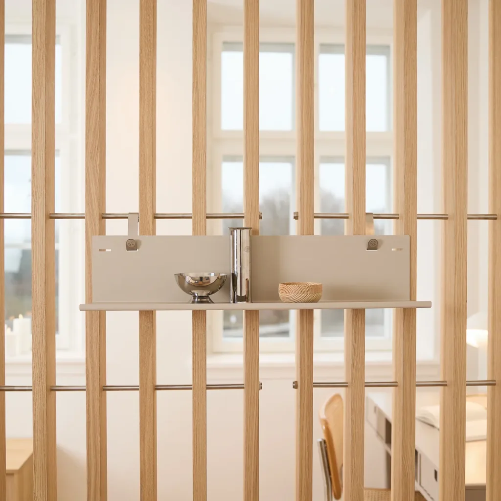 Divisione d’ambiente WoodUpp composta da listelli verticali in rovere chiaro con barre orizzontali metalliche, minimalista, e una piastra modulare WoodUpp color sabbia con mensola sospesa. Sulla mensola: ciotola in acciaio, vaso cilindrico lucido e piccola ciotola in legno, staffe metalliche visibili.