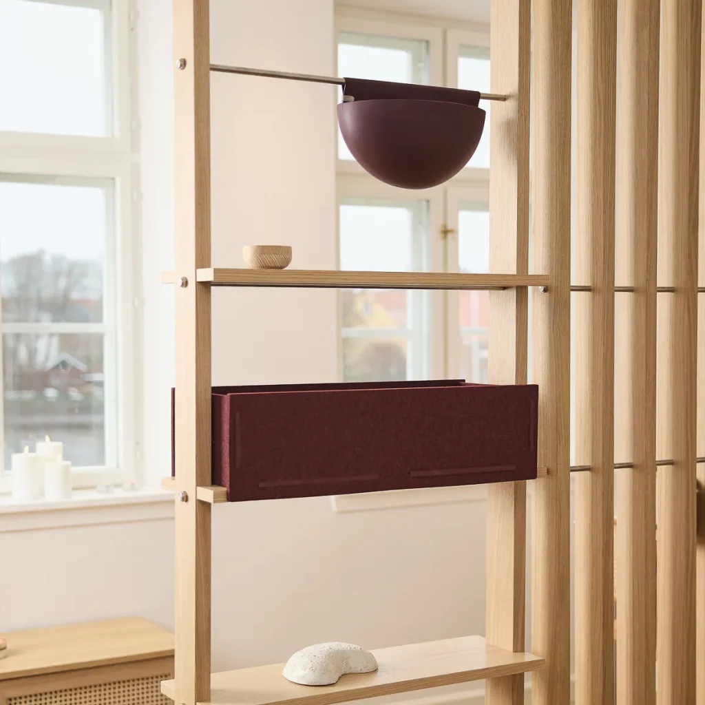Immagine di WoodUpp che mostra una struttura divisoria modulare in legno chiaro con ripiani, un contenitore organizer in feltro bordeaux montato su un ripiano e un vaso/portapiante sospeso semisferico nello stesso tono. Piccola ciotola di legno e pietra decorativa sui ripiani.
