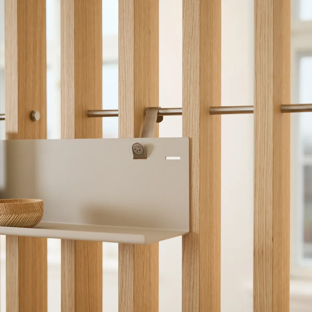 Immagine WoodUpp che mostra un divisorio a listelli in rovere chiaro con asta metallica e una mensola sospesa in metallo beige agganciata tramite gancio; sulla mensola un piccolo contenitore/bowl in legno intagliato. Dettaglio di finiture naturali e supporti metallici minimaliste ed eleganti.