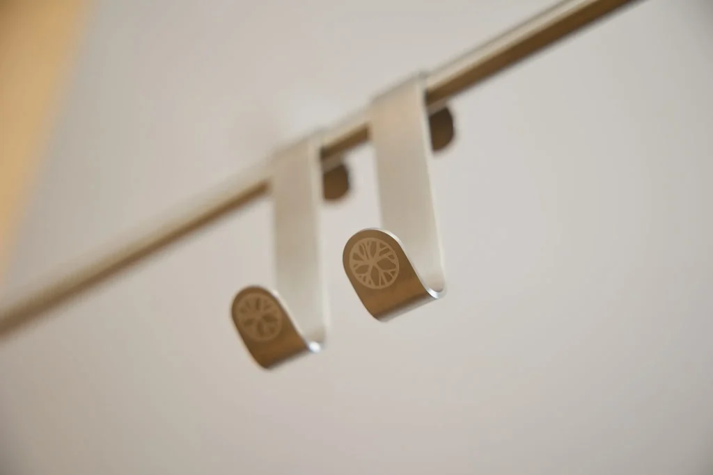Due ganci WoodUpp identici in metallo satinato, dal profilo sottile a S, appesi a un'asta orizzontale, con estremità ricurve per appendere oggetti. Sul lato frontale è visibile un'incisione circolare a forma di ruota, design minimalista e finitura opaca, sfondo neutro sfocato.