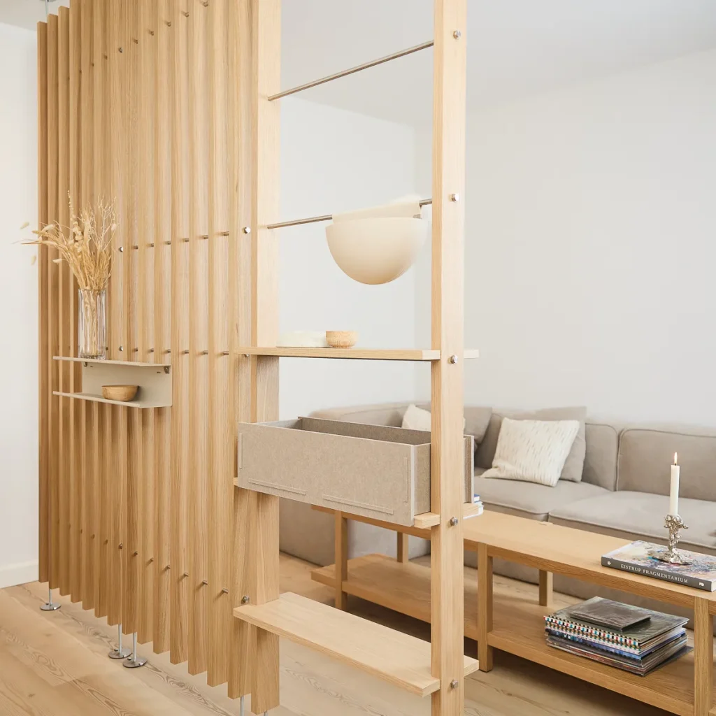 Parete divisoria modulare WoodUpp in legno chiaro con doghe verticali; include ripiani regolabili, mensola con vaso di vetro e ciotola, cassetto contenitore beige in feltro estraibile e barra metallica con sospensione a ciotola/lampada rotonda. Accoglie oggetti decorativi e separa il soggiorno.