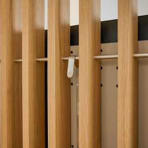 Pannello modulare WoodUpp con listelli verticali in legno di rovere chiaro, pannello posteriore forato e binario orizzontale in acciaio. Si vede un gancio appendiabiti WoodUpp in metallo satinato fissato al binario, dettaglio del design minimal e finiture naturali dall'estetica moderna.