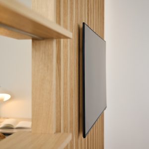 Modulo WoodUpp: pannello verticale a doghe in legno chiaro con mensole integrate e televisore piatto montato a filo. Si vedono listelli regolari in rovere chiaro, ripiani orizzontali e il supporto TV sottile che si incastra perfettamente nella struttura, dal design minimalista.