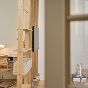 Pannello divisorio WoodUpp in rovere chiaro con struttura a listelli verticali e ripiani aperti, dotato di supporto porta TV sottile, mensola porta telecomandi metallica e scatola contenitore bianca montata. Scorcio di scrivania e sedia in paglia di Vienna sullo sfondo.