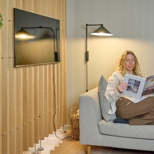 Interno con donna sul divano che legge; sul muro pannellatura verticale a listelli in legno WoodUpp, con supporti a pavimento in metallo e profilo per cavi visibile, TV sospesa e applique nera. Immagine mostra i pannelli WoodUpp e i loro accessori di fissaggio.