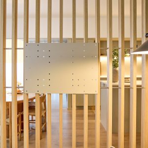 Separé a listelli in legno chiaro con installato il pannello di montaggio WoodUpp: una piastra metallica forata, fissata su due aste orizzontali in acciaio inserite tra le doghe. Sullo sfondo zona pranzo e cucina illuminate con arredi in legno dallo stile minimalista.