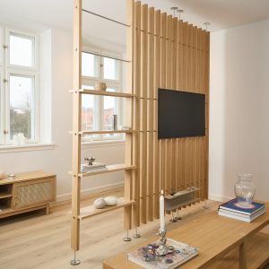 Parete divisoria WoodUpp in rovere chiaro con doghe verticali sospese fino al soffitto, supporto TV integrato e scaffalature laterali aperte regolabili su tubi metallici. Ambiente luminoso con mobili coordinati, oggetti decorativi e pavimento in legno chiaro, atmosfera minimal e nordica.