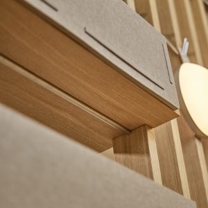 Dettaglio ravvicinato di un'installazione WoodUpp: pannelli fonoassorbenti in feltro grigio sovrapposti a una mensola e listelli in rovere chiaro WoodUpp; si vedono il bordo della mensola, il montante verticale e una lampada da parete ovale che illumina la texture del legno.