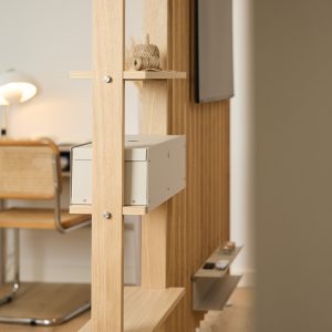 Divisorio modulare in rovere WoodUpp con struttura a doghe sottili, mensole incrociate e un contenitore metallico sospeso color grigio chiaro (cassettino) di WoodUpp; un rotolo di spago su mensola e una mensola inferiore con telecomando, sullo sfondo scrivania e sedia.